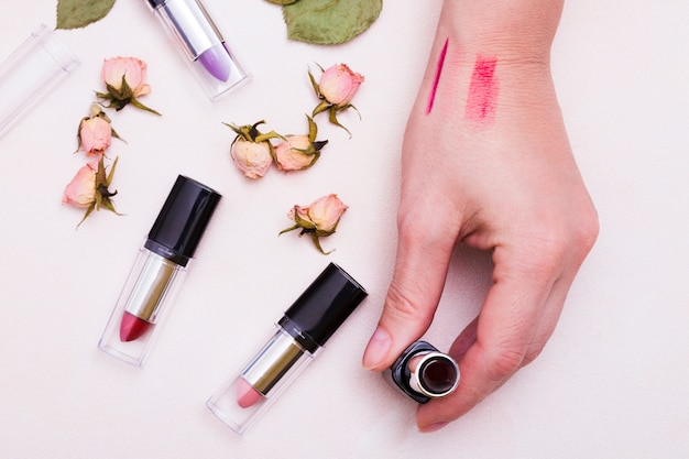 An elevated view of female hand holding lipstick with dried pink rose buds on white backdrop