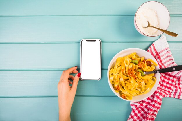 Elevated view of female's hand near smartphone and tagliatelle pasta