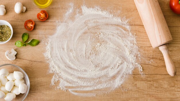 An elevated view of flour with tomatoes; mushroom; basil and mozzarella cheese on wooden table with rolling pin on table