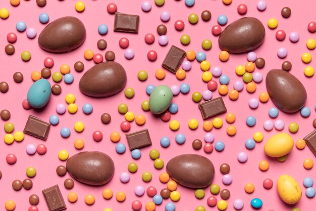 An elevated view of gem candies and chocolate easter eggs on pink background