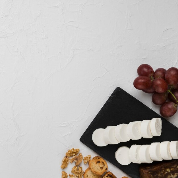Повышенный вид ломтик козьего сыра на сланцевой скале с виноградом; хлеб и орех на текстурированном фоне