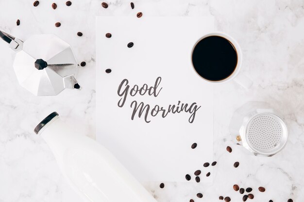 An elevated view of good morning text on paper; cafeteria coffee pot; coffee cup; milk bottle and coffee beans on marble backdrop