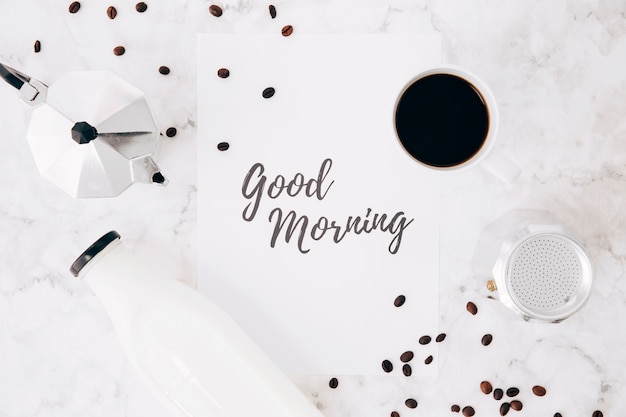 Поднятый вид текста доброго утра на бумаге; кафетерий кофейник; чашка кофе; бутылка молока и кофейные зерна на мраморном фоне