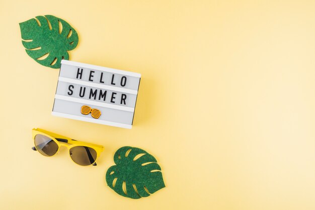 An elevated view of green artificial monstera leaves; sunglasses near the hello summer light box on colored background