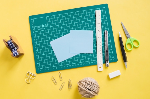 Elevated view of green cutting mat and stationeries on yellow background
