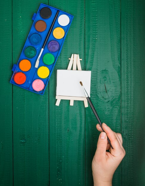 Elevated view of hand holding paint brush over mini easel and watercolor palette