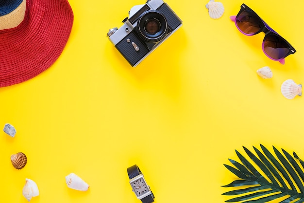 Elevated view of hat; camera; sunglasses; sea shells; wrist and palm leaf on yellow surface