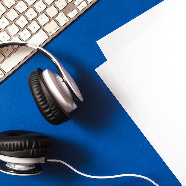 Elevated view of headphone; paper and keyboard on blue background