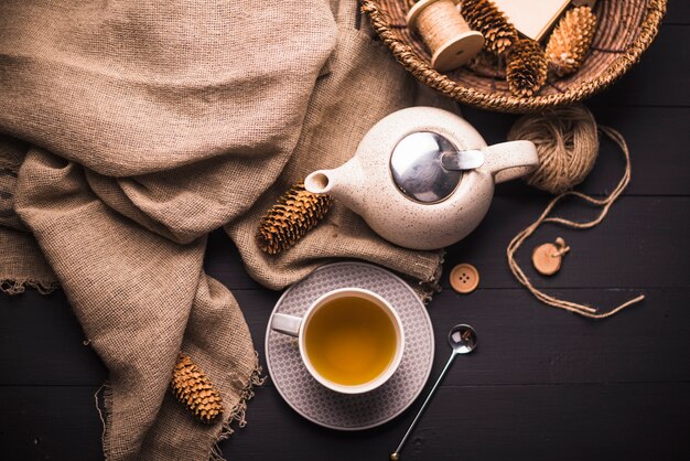 Elevated view of herbal tea; pinecone; teapot; sack; button; wicker basket and ball of yarn on table