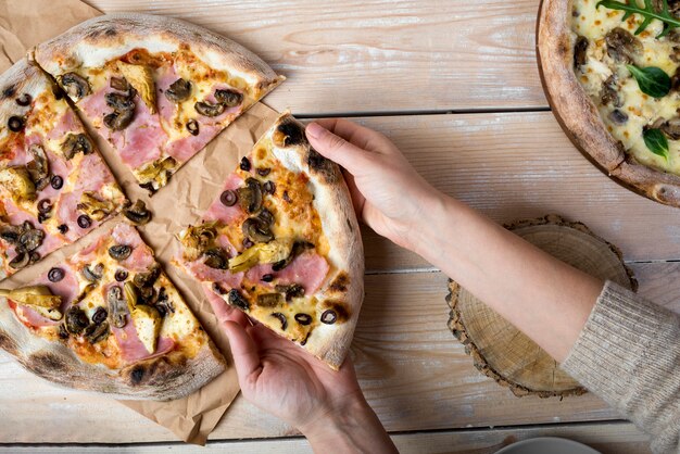 Elevated view of a human hand taking slice of pizza from brown paper over wooden table