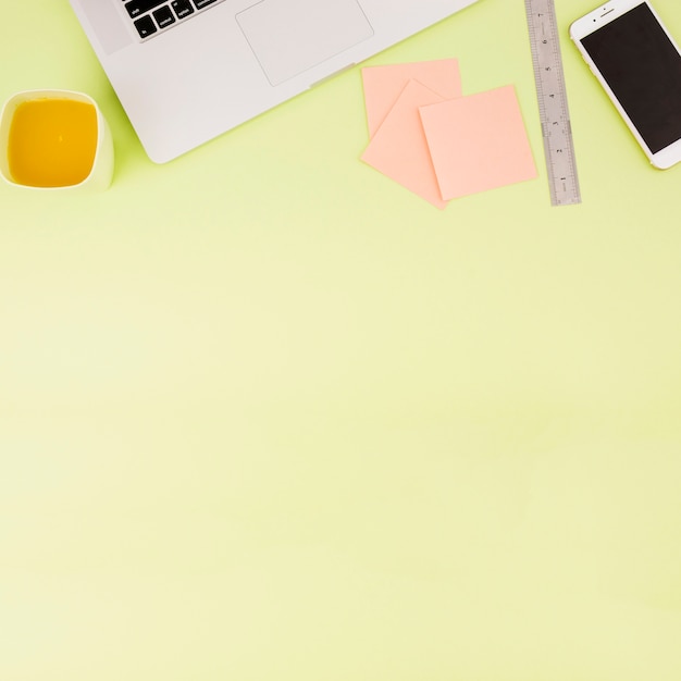 Elevated view of juice; laptop; smartphone; adhesive notes and ruler on colored backdrop