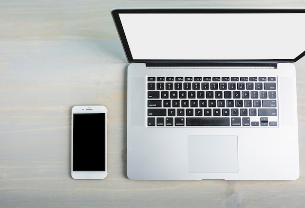 Elevated view of laptop with blank white screen and cellphone on wooden desk