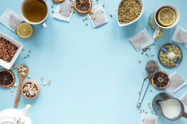 Elevated view of lemon tea; herbs; milk; strainer; dried chinese chrysanthemum flowers; teapot and teabags arranged on blue background