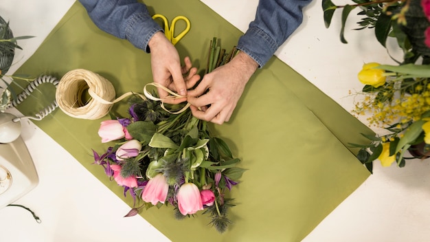 An elevated view of male tourist tying the flower bouquet with string on green paper over the desk