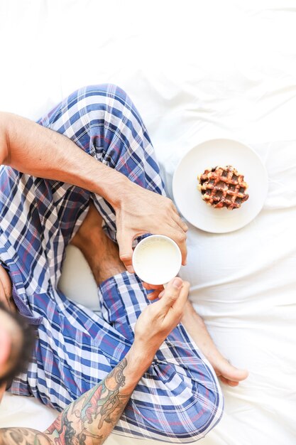 Foto gratuita punto di vista elevato dell'uomo con la tazza di latte e cialda che si siedono sul letto