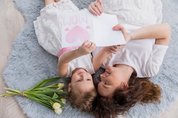 Elevated view of mother and daughter lying on blue fluffy carpet with flowers and greeting card