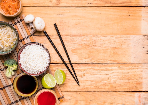 An elevated view of mushroom; sprout beans; rice; broccoli; lemon; and sauce with chopsticks against wooden desk