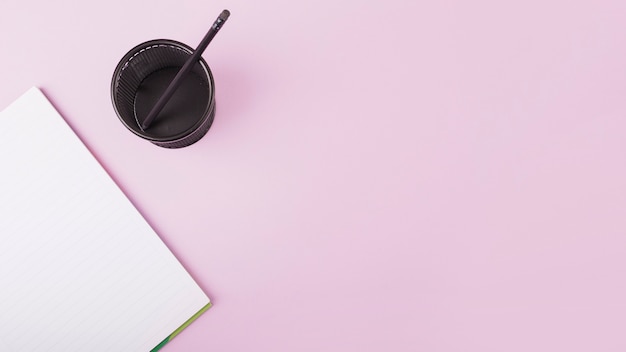 Elevated view of notebook and plastic cup with pencil on pink backdrop
