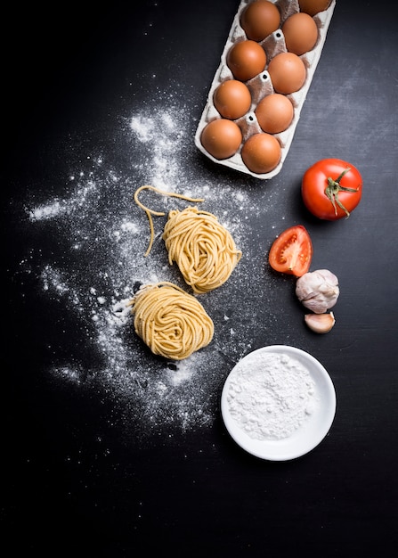 무료 사진 계란 판지를 가진 capellini 파스타의 높은 전망; 육즙 토마토; 마늘과 카운터 위에 밀가루의 그릇