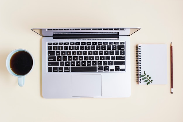 Elevated view of opened laptop; tea cup; spiral notepad and pencil on beige background