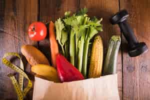 Free photo elevated view of paper bag with vegetables near dumbbell and measuring tape