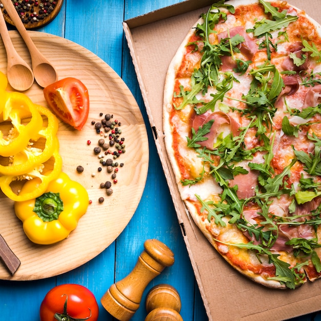 Elevated view of pepperoni pizza in cardboard box with spices; peppermill and vegetables over blue wooden table