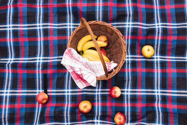 An elevated view of picnic basket with bananas and apples on blanket