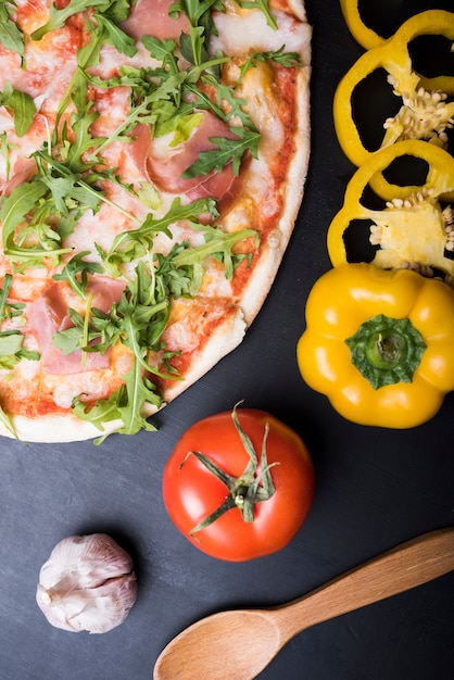 Free photo elevated view of pizza with bacon and arugula leaves near sliced yellow bell pepper; garlic bulb; tomato and wooden spoon