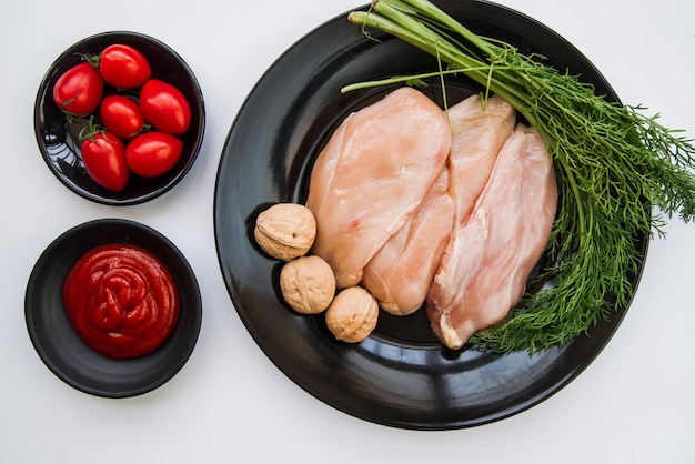 Elevated view of raw chicken; fresh dill; walnut; tomato and sauce on white background