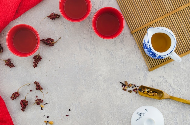 An elevated view of red ceramic cups and herbal tea in pitcher on textured background