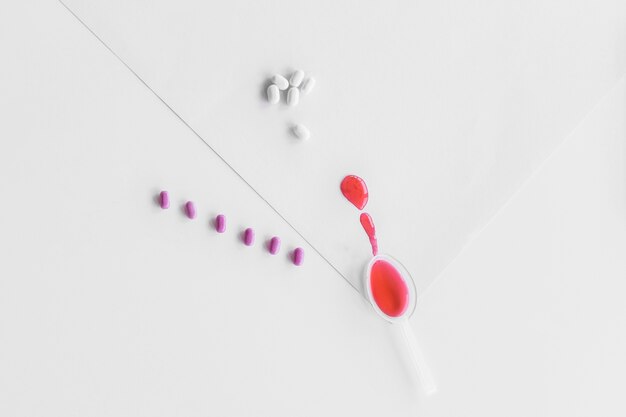 Elevated view of red syrup in small spoon with row of purple and white pills