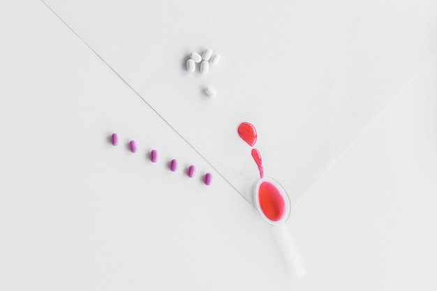 Elevated view of red syrup in small spoon with row of purple and white pills