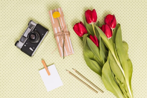 Elevated view of retro camera; blank paper; pencils; gift box and bouquet of tulip flowers over yellow background