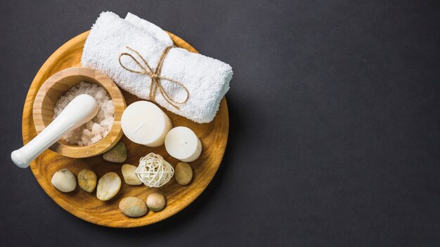 Elevated view of salt; towel; candles and spa stones on wooden plate over black backdrop