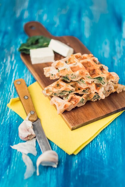 Elevated view of salty waffles on wooden board with cheese; knife and garlic cloves over blue textured background
