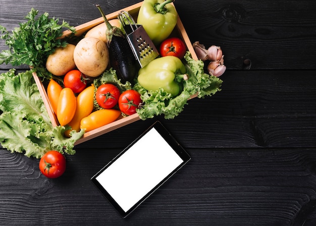 Elevated view of smartphone near vegetables in container on black wooden surface