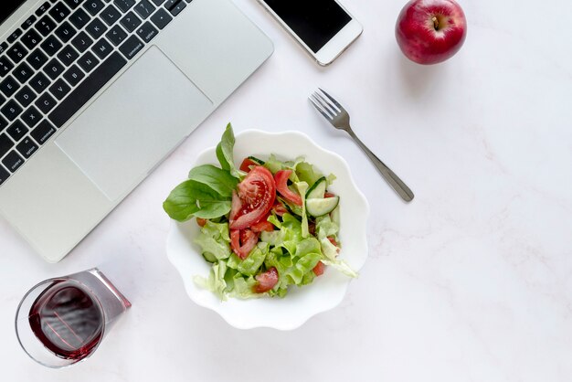 Elevated view of soft drink; bowl of salad; apple and fork near electronic devices over white background