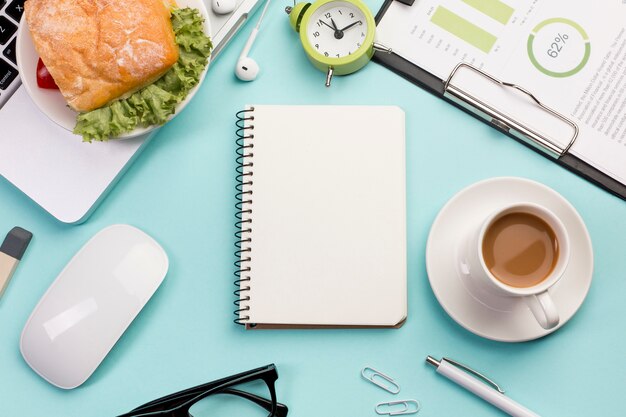 An elevated view of spiral notepad,breakfast,mouse and laptop on office desk