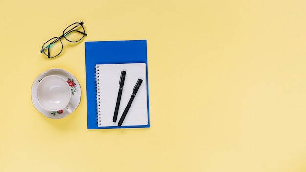 Elevated view of spiral notepad; cup and eyeglasses on yellow background