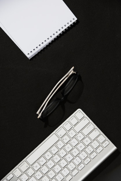 An elevated view of spiral notepad; eyeglasses and keyboard on black desk