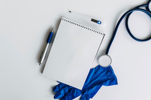 Elevated view of spiral notepad; pen; stethoscope; thermometer and gloves on white backdrop