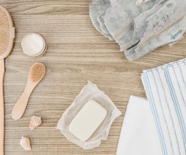 Elevated view of sponge; seashell; soap; brush; towel and moisturizing cream on wooden background