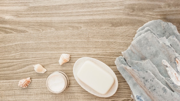 Elevated view of sponge; seashell; soap and moisturizing cream on wooden surface