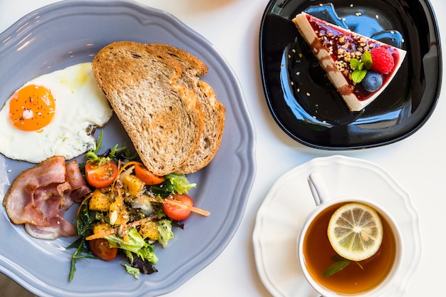 An elevated view of tasty cheesecake; lemon tea cup; toast; salad; fried egg and bacon on gray plate over white background