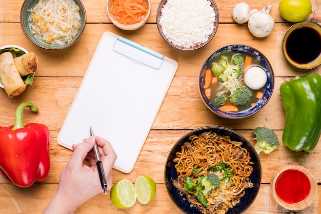 Поднятый вид тайской еды с человеком, пишущим в буфер обмена с ручкой
