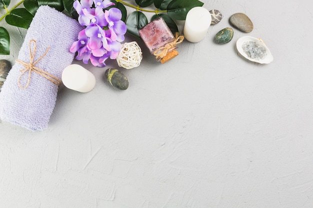 Elevated view of towel; candles; scrub bottle; flowers and spa stones on grey backdrop