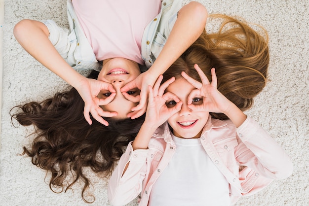 Free photo an elevated view of a two female friend lying on carpet doing ok gesture like binocular looking up