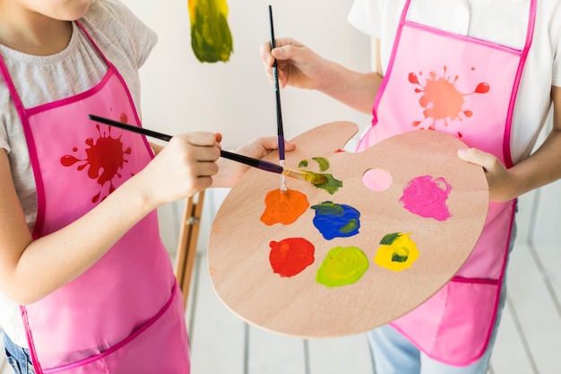 An elevated view of two girls in same pink apron mixing the paint on wooden palette with paint brush