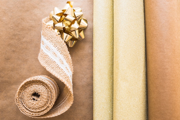 Elevated view of weaving ribbon and golden bow with gift paper on brown paper