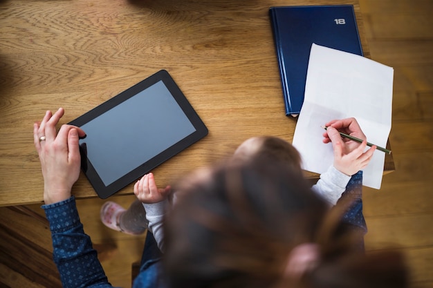 Elevated view of woman writing notes on paper while using digital tablet